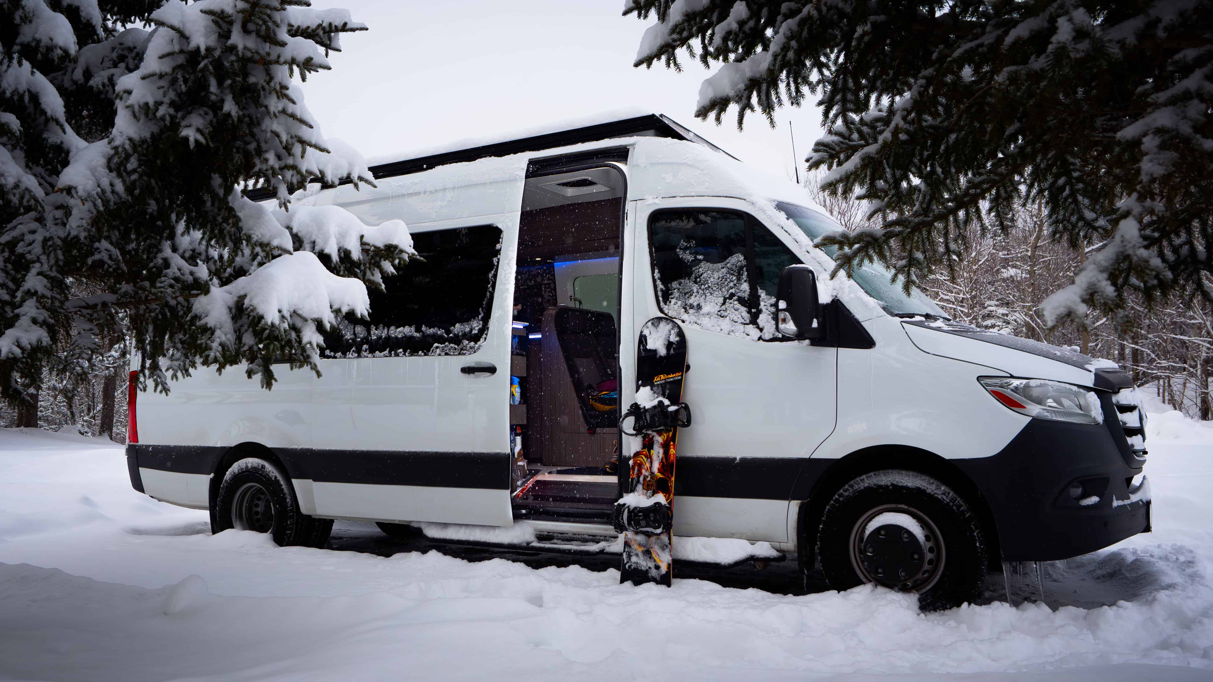 Fourgon aménagé Icon Vans blanc parked in forêt enneigée with neigeboard près de la porte